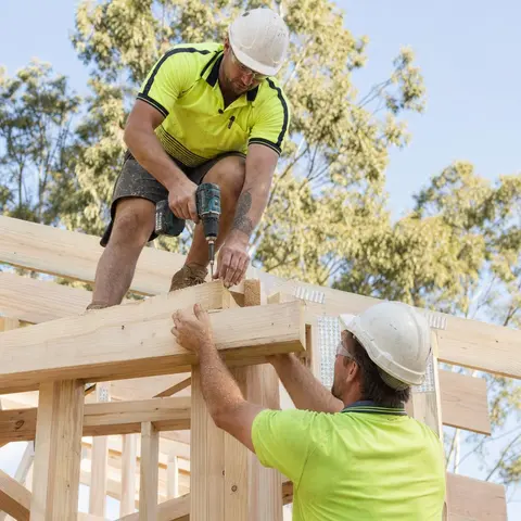 Illustrative image of two tradies fastening a timber house frame in a Queensland-style setting.